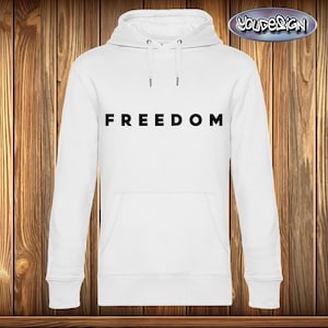 Könnte beinhalten: Weißer Kapuzenpullover mit dem Wort „FREEDOM“ in Schwarz auf der Brust. Er hat eine Kapuze mit Kordelzug und eine Vordertasche. Der Hintergrund ist eine Holzwand.