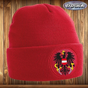 Könnte beinhalten: Rote Strickmütze mit einem schwarzen und gelben gestickten Aufnäher, der einen schwarzen Adler mit einem rot-weißen Schild und einer Krone auf dem Kopf zeigt.