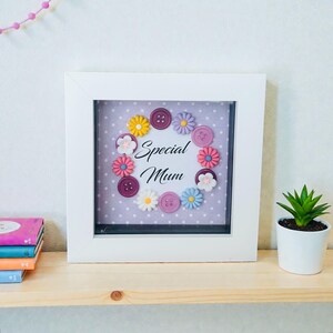 以下が含まれることがあります： 「Special Mum」の文字が書かれた白いフレームのアートワーク。カラフルな花の形をした装飾とボタンで囲まれています。背景は白のドットが入った薄紫です。右側には小さな鉢植えの植物があります。