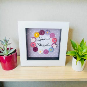Può includere: Un'opera d'arte con cornice bianca con la scritta "Special Daughter" circondata da decorazioni a forma di fiori colorati e bottoni. Lo sfondo è lilla chiaro con pois bianchi. L'opera è esposta su una mensola di legno con piante in vaso.