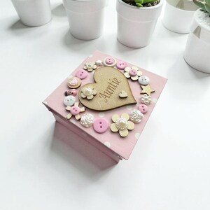 May include: A pink square gift box with white polka dots, adorned with a wooden heart that says "Auntie". The heart is surrounded by decorative buttons and wooden flowers. The box is on a white surface.
