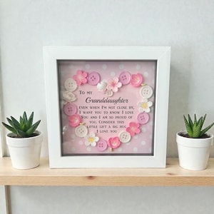 Puede incluir: Una obra de arte enmarcada en blanco con un diseño en forma de corazón hecho de botones y flores rosas y blancas. Se muestra el texto "TO MY Granddaughter", junto con un mensaje de amor. Dos pequeñas suculentas en macetas flanquean el marco.