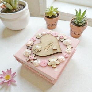 Peut inclure: Un coffret rose en forme de carré, décoré de fleurs en bois, de boutons et d'un cœur en bois avec le mot "Nan". La boîte est entourée de petites plantes succulentes en pot et d'une seule fleur rose.