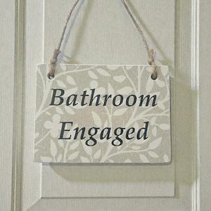 May include: A rectangular wooden sign with the words "Bathroom Engaged" in dark grey lettering. The sign has a light grey background with a white floral pattern. It hangs from a natural twine string.