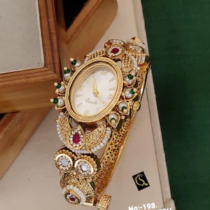 May include: A gold-tone analog watch with an oval face, white dial, and gold hands. The watch features an ornate, filigree-style band decorated with small white pearls, green beads, and red stones. Text on the image reads: No:-198. ANTIQUE WATCH D:-RM - 0022.