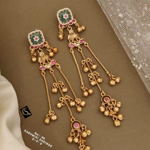 Könnte beinhalten: Ein Paar goldfarbene Ohrhänger mit quadratischen grünen Zentren, weißen Steinakzenten, Elefantenmotiven und mehreren glockenförmigen Anhängern. Die Ohrringe enthalten rosa Steindetails und sind beschriftet mit No: 46, KASHMIRI EARINGS, D:-P- 6.
