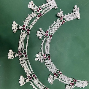 May include: A pair of silver-toned anklets featuring multiple delicate chains, accented with floral-shaped motifs containing small red and green stones. Each anklet is adorned with dangling silver-toned charms and small bells along the length.