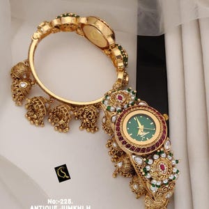 Könnte beinhalten: Eine goldfarbene Armbanduhr mit rundem grünem Zifferblatt, einer mit roten Steinen besetzten Lünette und baumelnden glockenförmigen Anhängern. Das Armband ist mit weißen Perlen, grünen Perlen und klaren Steinen verziert. Text: No:-225. ANTIQUE JUMKHI H 1- PICS WATCH D:-RM - 0024