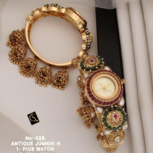 Puede incluir: Un reloj tipo brazalete dorado con esfera redonda y bisel incrustado con piedras rojas y verdes. El reloj está adornado con múltiples dijes dorados colgantes en forma de campana. El texto dice: No:-225. ANTIQUE JUMKHI H 1- PICS WATCH D:-RM - 0024.