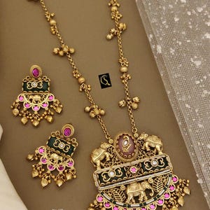 May include: A gold-toned heritage pendant set featuring a necklace and matching earrings. The necklace has a large pendant with elephant and floral designs, accented with pink and green gemstones. The earrings mirror the pendant's design. The set is ornate and detailed.