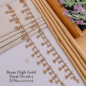 Könnte beinhalten: Ein Paar goldfarbene Metall-Fußkettchen mit Kettendesign, hängenden Blumenanhängern, kleinen Glöckchen sowie roten und grünen Steinen. Der Text "Braas High Gold Payal No.96.5 D.No.000105" ist unten links in Schwarz auf das Bild gedruckt.