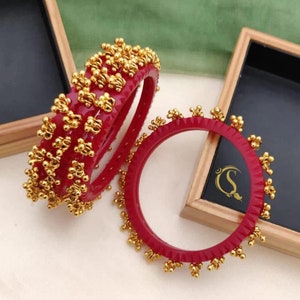 May include: Set of four red bangles with gold accents. The bangles are decorated with small gold balls and have a textured surface. The bangles are arranged in a circle, with the largest bangle on the bottom and the smallest bangle on top. The bangles are in a wooden box with a black interior.