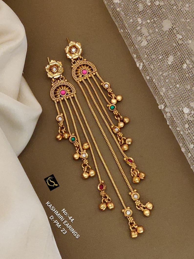 May include: Gold-toned Kashmiri earrings with floral and half-moon designs. The earrings feature dangling chains with small beads and colorful accents of pink, green, and white. The text on the image reads "Kashmiri Earrings".