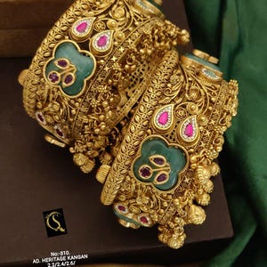 Può includere: Due braccialetti dorati con intricati motivi floreali e geometrici. Ogni braccialetto presenta dettagli verde smeraldo e rosso rubino. Il testo sull'immagine recita: "No:-310. AD. HERITAGE KANGAN 2.2/2.4/2.6/ D:-1058."