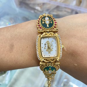 Op de afbeelding: Een goudkleurig analoog horloge met een rechthoekige witte wijzerplaat met een gouden godheidillustratie in het midden. De rand is bezet met heldere stenen en de armband bevat sierlijke gouden schakels met donkergroen email en kleine rode steentjes.