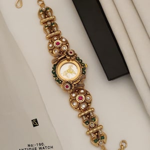 May include: A gold-toned watch with a round face featuring a horse graphic. The band is composed of ornate metal links decorated with small white stones, green beads, and central red stones. Text on the image reads: No:-190. ANTIQUE WATCH D-RM-0019-W.
