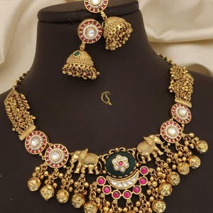 May include: A gold-toned necklace and jhumka earring set featuring elephant motifs, circular accents with white stones, and red stone inlays. The necklace includes dangling gold beads and a central dark green circular pendant. Text reads: No:-235. HERITAGE NECKLACE D:-1018