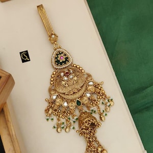 May include: An elaborate gold-toned hair ornament with detailed patterns. It includes a triangular top with a floral design, a large circular central piece, and hanging bell-shaped components. The text "No-65. HERITAGE JUDA D-SK-8009" is visible.