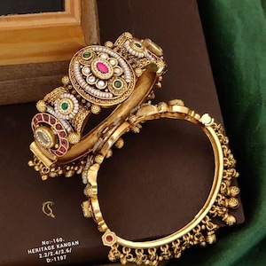 Puede incluir: Dos brazaletes dorados con detalles intrincados. Uno presenta un diseño circular grande con acentos rojos, verdes y blancos. El otro tiene una serie de pequeñas cuentas doradas colgantes. El texto "No:-160. HERITAGE KANGAN 2.2/2.4/2.6/ D:-1197" es visible.