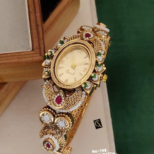 May include: An oval-faced gold-tone quartz watch with an ornate metal band featuring intricate filigree, embedded white pearls, and red and green stones. The watch face is gold with gold hands and the text No:-198. ANTIQUE WATCH D:-RM - 0022.