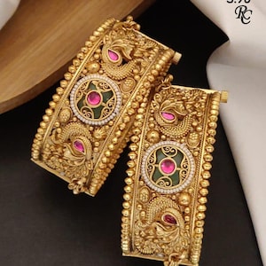 Peut inclure: Deux bracelets dorés ornés de motifs de paon en relief, de bords perlés et d'un médaillon central circulaire avec une incrustation verte et une pierre rose. Le texte "Size 22.24 Heritage Patla S.98" et un logo sont visibles en haut à droite.
