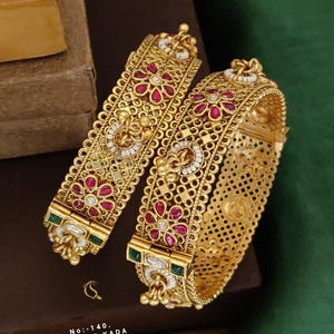 Op de afbeelding: Twee goudkleurige scharnierende kada-armbanden met ingewikkeld filigraan metaalwerk, ingelegd met rode bloemvormige stenen, kleine witte kralen en groene rechthoekige accenten. De tekst op de afbeelding luidt No: 140, ANTIQUE KADA, 2.2/2.4/2.6, D: AN-1216.