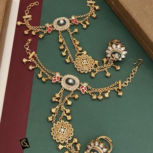 Könnte beinhalten: Ein Paar goldfarbene Handketten mit filigranen Details, dunkelblauen runden Elementen und roten Akzenten. Jedes Stück besteht aus einem Ring, der durch zarte Ketten mit kleinen Anhängern verbunden ist. Text: No:-254. HERITAGE PANJA JODI D:-M - 5022.