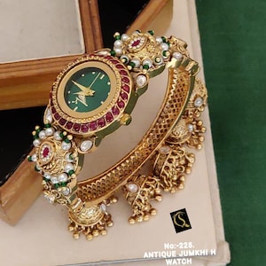 May include: A gold-toned bangle watch featuring a round green dial with gold hands, surrounded by a bezel of red stones. The band is decorated with white pearls, green beads, and dangling gold bell-shaped charms. Text reads: No:-225. ANTIQUE JUMKHI H WATCH D:-RM-0024.