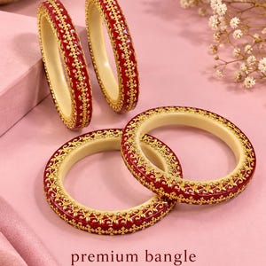 Könnte beinhalten: Vier kreisförmige Armreifen mit cremefarbener Innenseite und einer tiefroten Außenseite, die mit komplizierten goldfarbenen Filigranmustern verziert ist. Der Text "premium bangle" und "SG_NO_34" ist unten dunkelrot aufgedruckt.