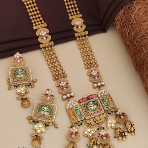 Puede incluir: Un collar largo de tono dorado y un juego de pendientes cuadrados a juego con centros de esmalte verde, piedras rosas, cuentas blancas y dijes dorados colgantes. El texto D.No-ABP-44 Brass High Gold Designer Long Set No.190 está impreso en la parte inferior izquierda.