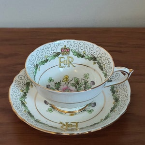 Op de afbeelding: Een witte theekop en schotel set met gouden rand, met een bloemen- en klaverontwerp. De theekop en schotel hebben de letters "ER" en een kroon. De rand van de theekop heeft een decoratief patroon.