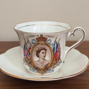 Op de afbeelding: Een witte theekop en schotel met gouden rand. De theekop toont een portret van een gekroonde vrouw, vlaggen en de tekst "June 2 1953". De schotel heeft een geschulpte rand.