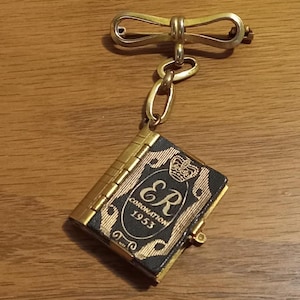 Op de afbeelding: Een gouden broche in de vorm van een strik met een ketting die is bevestigd aan een klein zwart boek met gouden letters die "Coronation 1953" en "ER" lezen met een kroon boven de letters.