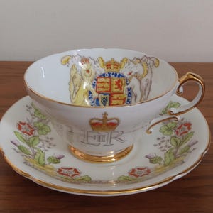Op de afbeelding: Een witte theekop en schotel set met gouden rand. De theekop heeft een koninklijk wapen en de initialen "ER" onder een kroon. De schotel is versierd met bloemen- en bladpatronen.