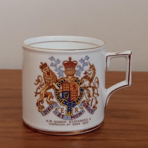 Op de afbeelding: Witte keramische mok met een gouden rand en oor, met een koninklijk wapen met een leeuw, een eenhoorn en een kroon. De mok heeft de tekst "H.M. Queen Elizabeth II Crowned 2nd June 1953".
