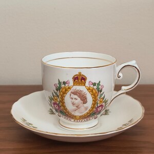 Op de afbeelding: Een wit theekopje en schoteltje met een gouden rand. Het theekopje heeft een portret van koningin Elizabeth II met een kroon en een bloemenontwerp. De tekst "Queen Elizabeth II" staat gedrukt op de onderkant van het theekopje.