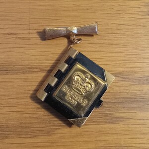 Op de afbeelding: Een gouden boekvormige broche met een zwarte emaille-deksel. De broche heeft een kroon en de tekst "Elizabeth II 1953".