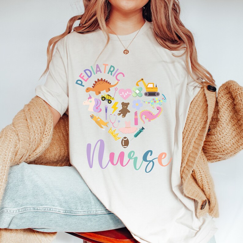 May include: A white t-shirt with the text "Pediatric Nurse" in rainbow colours. A heart shape is formed by various colourful cartoon images of toys and objects associated with children, such as a dinosaur, a teddy bear, a crown, a stethoscope, a toy truck, a unicorn, a lightning bolt, a bandage, a heart monitor, a flower, a dragon, a toy airplane, a crown, a football, and a syringe.