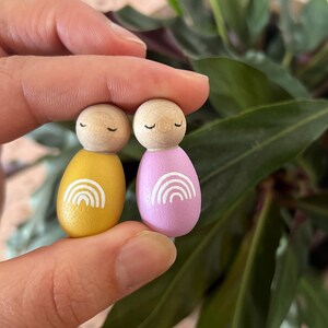 May include: Two wooden peg dolls, one yellow and one pink, each with a white rainbow design. The dolls have wooden heads with closed eyes. The dolls are held in a hand, with a green leafy background.