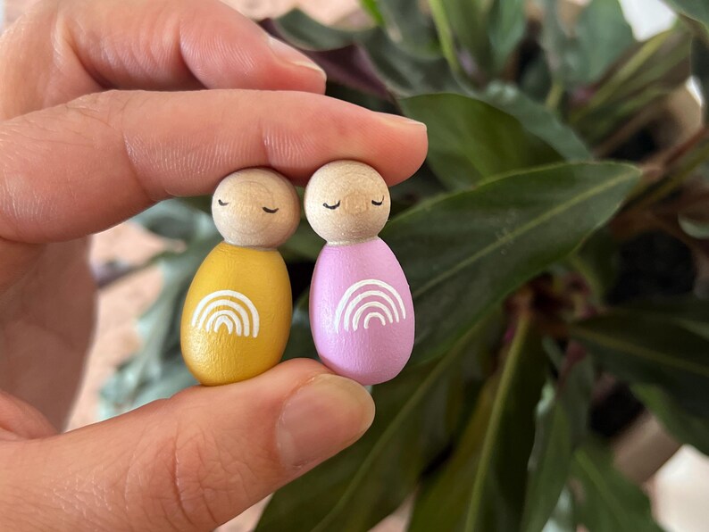 May include: Two wooden peg dolls, one yellow and one pink, each with a white rainbow design. The dolls have wooden heads with closed eyes. The dolls are held in a hand, with a green leafy background.