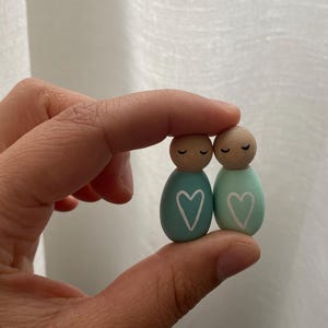 May include: Two wooden peg dolls, each with a painted teal body and a wooden head. The dolls have closed eyes and a white heart painted on the front. The dolls are held together.