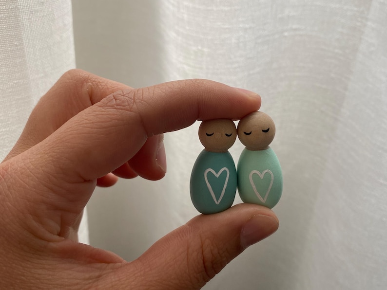 May include: Two wooden peg dolls, each with a painted teal body and a wooden head. The dolls have closed eyes and a white heart painted on the front. The dolls are held together.