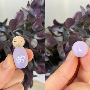 May include: A wooden peg doll with a light purple body and a butterfly design. The doll has a wooden head with closed eyes. A light purple ball with the letter "L" is also shown. The background features purple flowers.