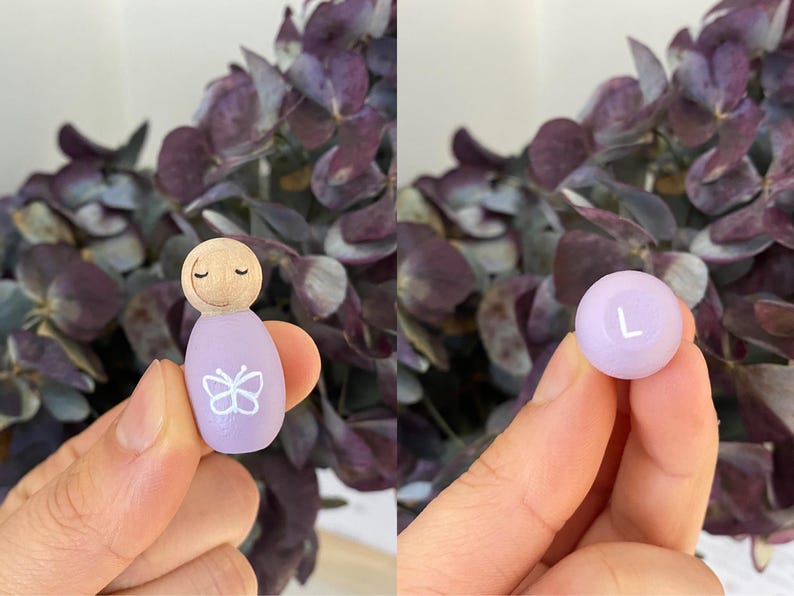 May include: A wooden peg doll with a light purple body and a butterfly design. The doll has a wooden head with closed eyes. A light purple ball with the letter "L" is also shown. The background features purple flowers.