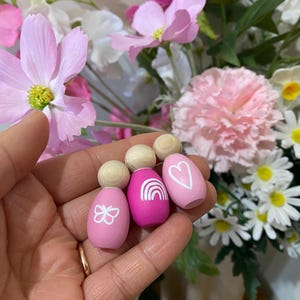 May include: Wooden peg dolls with painted designs. The dolls are pink and magenta, with a butterfly, rainbow, and heart design. The dolls have a wooden ball for a head. The background is a floral arrangement with pink and white flowers.