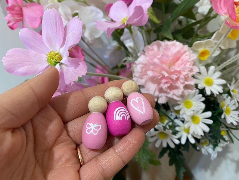 May include: Wooden peg dolls with painted designs. The dolls are pink and magenta, with a butterfly, rainbow, and heart design. The dolls have a wooden ball for a head. The background is a floral arrangement with pink and white flowers.