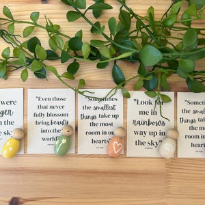 May include: Five small wooden peg dolls with inspirational quotes on cards. The dolls are painted in various colours, including yellow, green, orange, white, and purple, and feature simple designs like stars, flowers, a heart, a rainbow, and a butterfly. Greenery is arranged above.