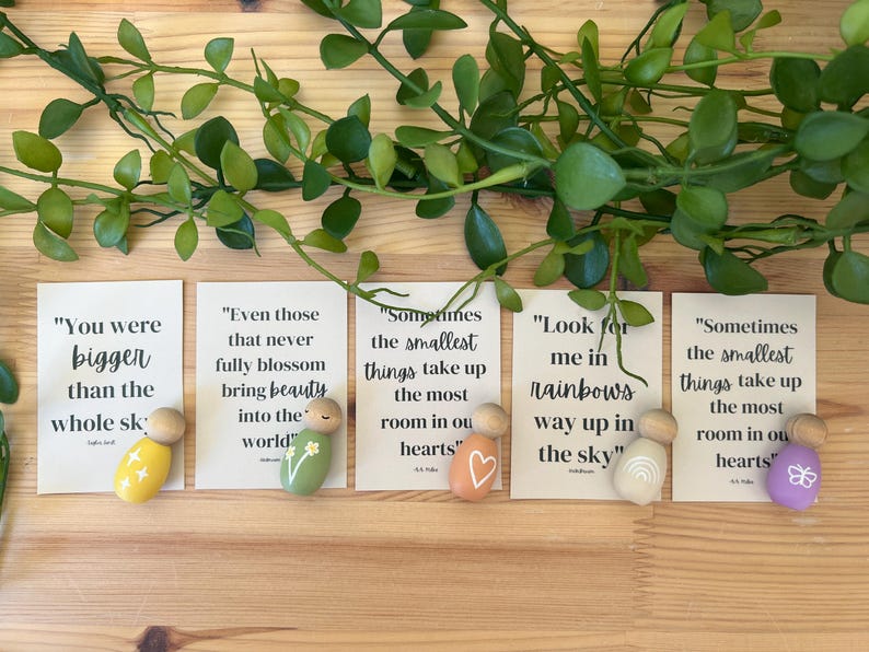 May include: Five small wooden peg dolls with inspirational quotes on cards. The dolls are painted in various colours, including yellow, green, orange, white, and purple, and feature simple designs like stars, flowers, a heart, a rainbow, and a butterfly. Greenery is arranged above.