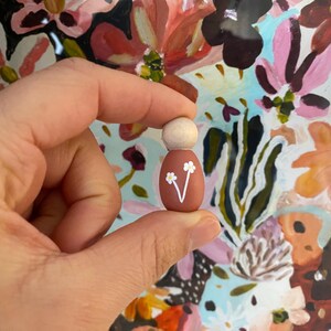 May include: A small wooden peg doll with a light brown head and a terracotta body. The body is decorated with a white floral design. The doll is held in a person's hand against a colourful floral background.