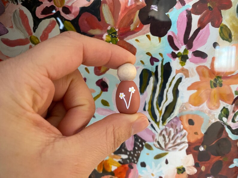 May include: A small wooden peg doll with a light brown head and a terracotta body. The body is decorated with a white floral design. The doll is held in a person's hand against a colourful floral background.